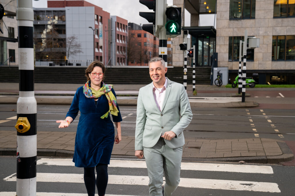 Sanne Boswinkel en Lodewijk van Noort steken over op een zebrapad, bij een groen voetgangerslicht, en kijken in de camera.