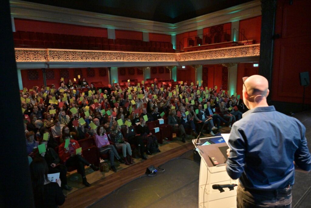 Dio stelt een vraag aan het publiek. Aanwezigen houden als antwoord een groen, oranje of rood blaadje in de lucht.