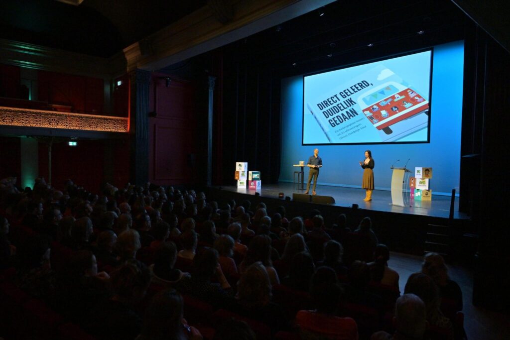 Volle zaal in theater Diligentia tijdens de boekpresentatie van 'Direct geleerd, duidelijk gedaan'. Op het podium dagvoorzitters Dio van der Veen en Talissa Oude Bennink. Met achter hen een groot scherm, met daarop de cover van het boek.