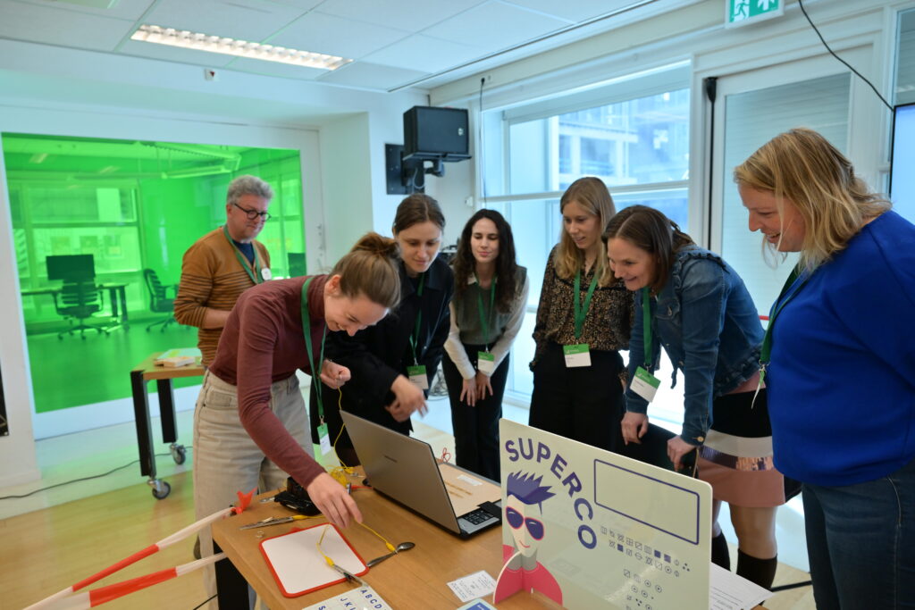 Lachende en serieuze gezichten van een team van 7 mensen rond een tafel met laptop en attributen (kabels, borden, een lepel) om een opdracht te voltooien