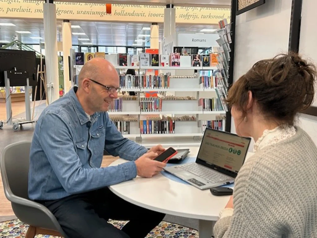 Digi-inloop van het Digi-Taalhuis: man met telefoon in zijn hand zit aan tafel bij een vrouw. Laptop op tafel. En rijen met boeken van de bibliotheek op de achtergrond.