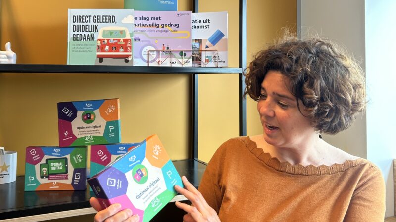 Persoon staat voor een boekenkast met Optimaal Digitaal spellen en heeft in haar handen de 4e druk van het spel