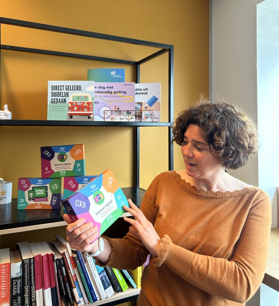 Persoon staat voor een boekenkast met Optimaal Digitaal spellen en heeft in haar handen de 4e druk van het spel