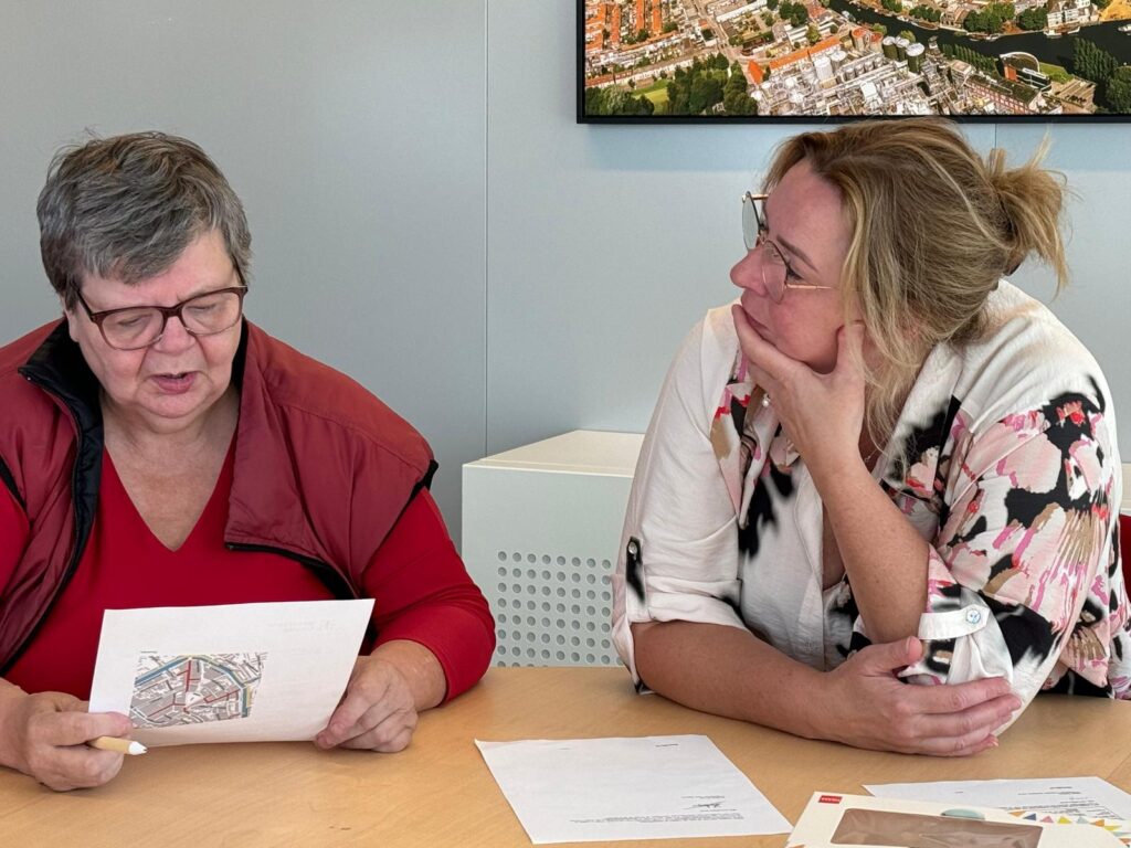 Foto van een taalambassadeur en Anita. Ze zitten aan een tafel en de taalambassadeur bekijkt een flyer.